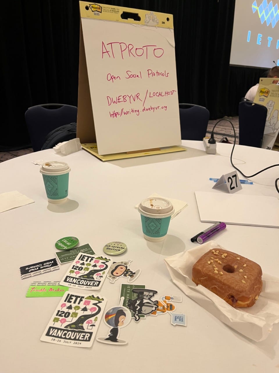 Conference table with various IETF and Github stickers, a large donut and coffees, and a sign "ATPROTO Social Protocols"